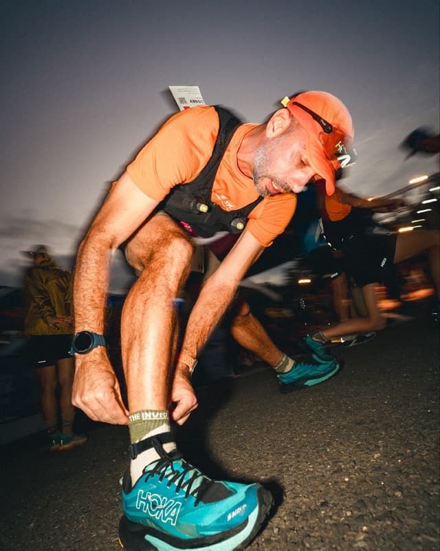 Male runner in orange adjusting his turquoise Hoka shoe, wearing a headlamp and hydration vest at night.