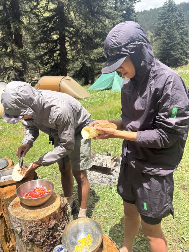 Two people prepare tacos at an outdoor campsite, using tree stumps as a table.