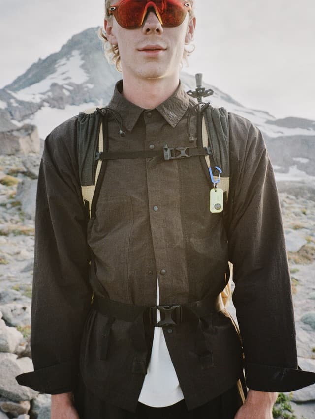 A man wearing red sunglasses, a dark shirt, and a backpack, with a snowy mountain in the background.