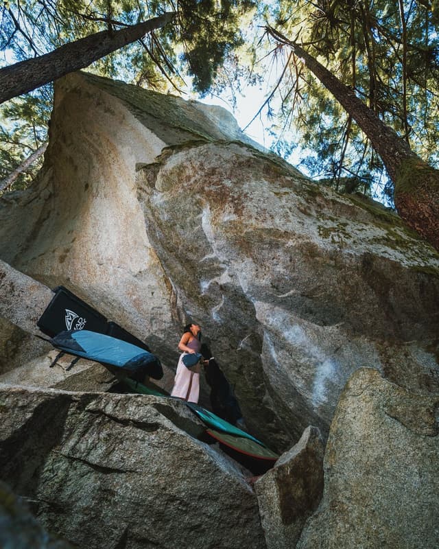 A woman stands among large boulders with bouldering crash pads in a forest, looking up at the rock face.
