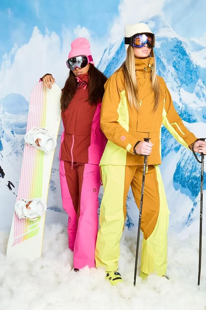 Two women in colorful ski and snowboard attire, one holding a striped snowboard and the other ski poles, against a mountain backdrop.