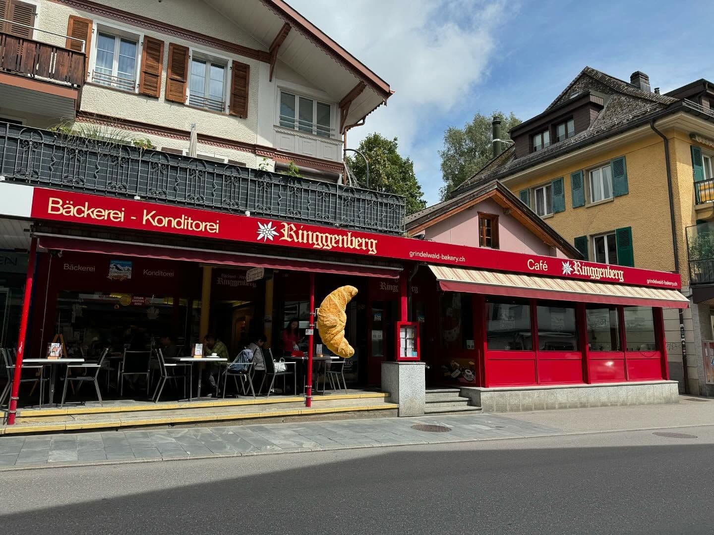 A red-fronted bakery and cafe named "Ringgenberg" with outdoor seating and a large croissant sign.