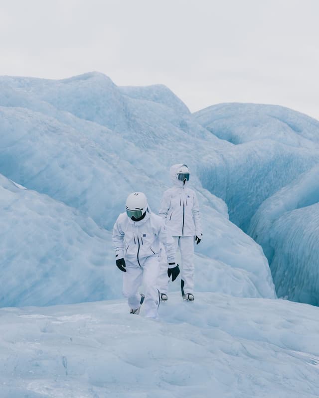 Two individuals in white winter suits and helmets walk on a blue glacier.