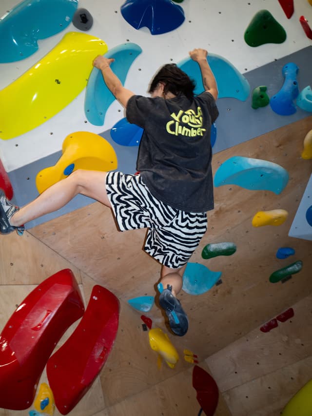 A person wearing a "Young Climber" t-shirt and zebra shorts bouldering on a colorful climbing wall.