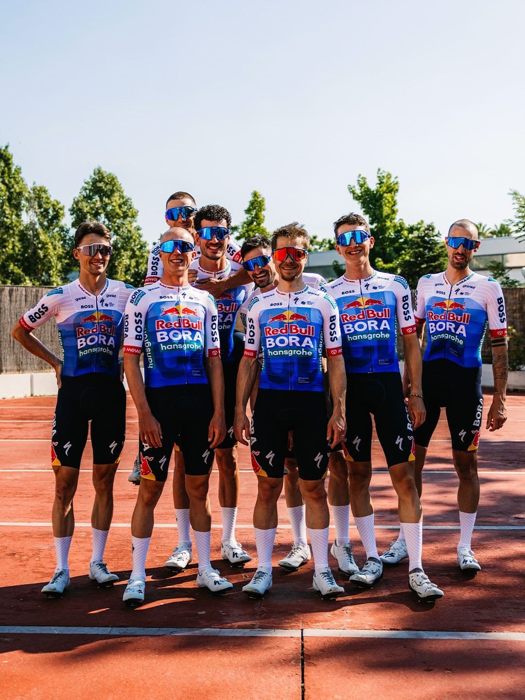 A group of smiling male cyclists in Red Bull Bora Hansgrohe team uniforms and sunglasses, posing on a red outdoor court.