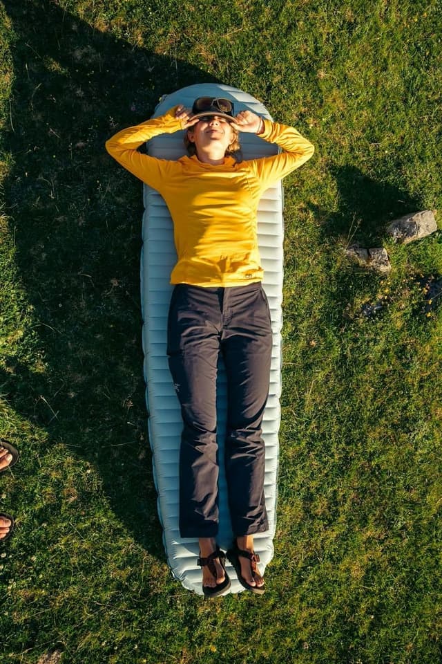 A person wearing a yellow shirt and dark pants lies on a sleeping pad in sunny grass, hands by their head.