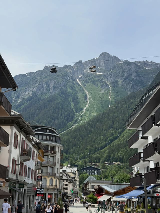 A busy street in a mountain town with buildings, leading towards a large, forested mountain, with shoes hanging from wires overhead.