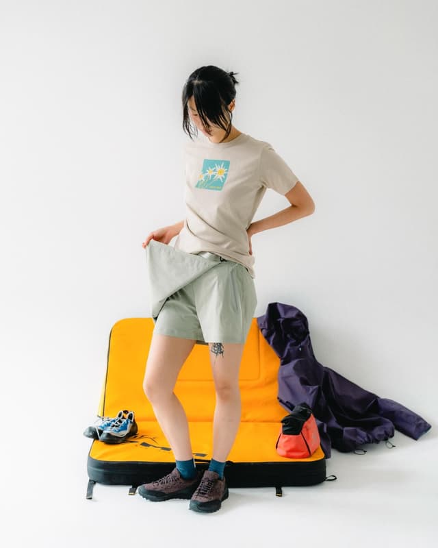 A person in a beige t-shirt with a flower graphic, green shorts, and trail shoes stands next to an orange bouldering mat with a purple jacket, chalk bag, and blue shoes.