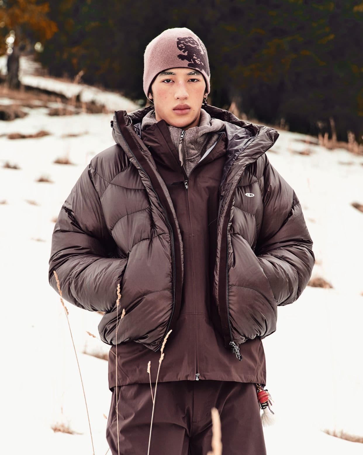 A person in a brown puffer jacket and patterned beanie stands in a snowy field.