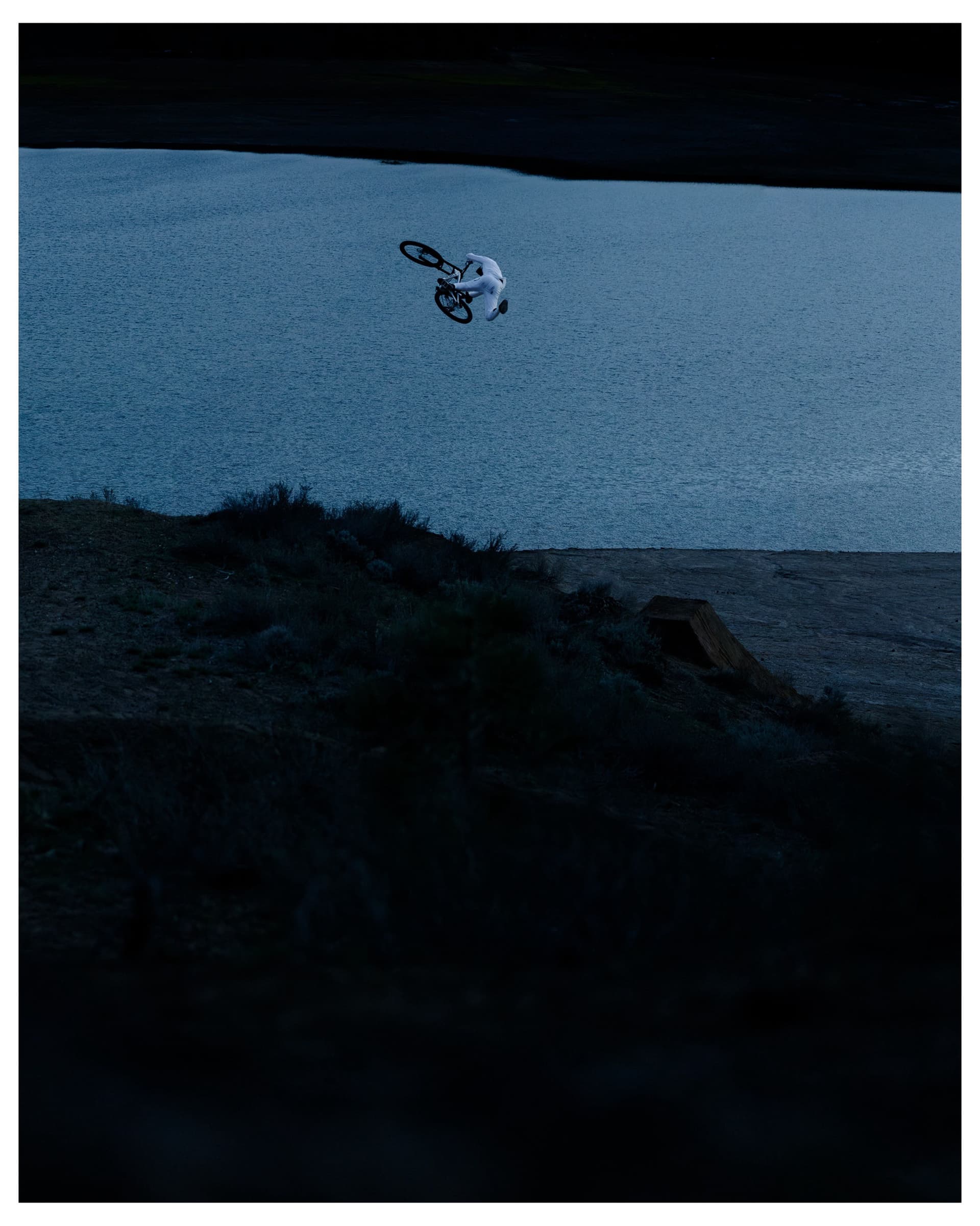A person on a BMX bike jumps high over a dark blue lake, with a grassy bank and dirt ramp visible.
