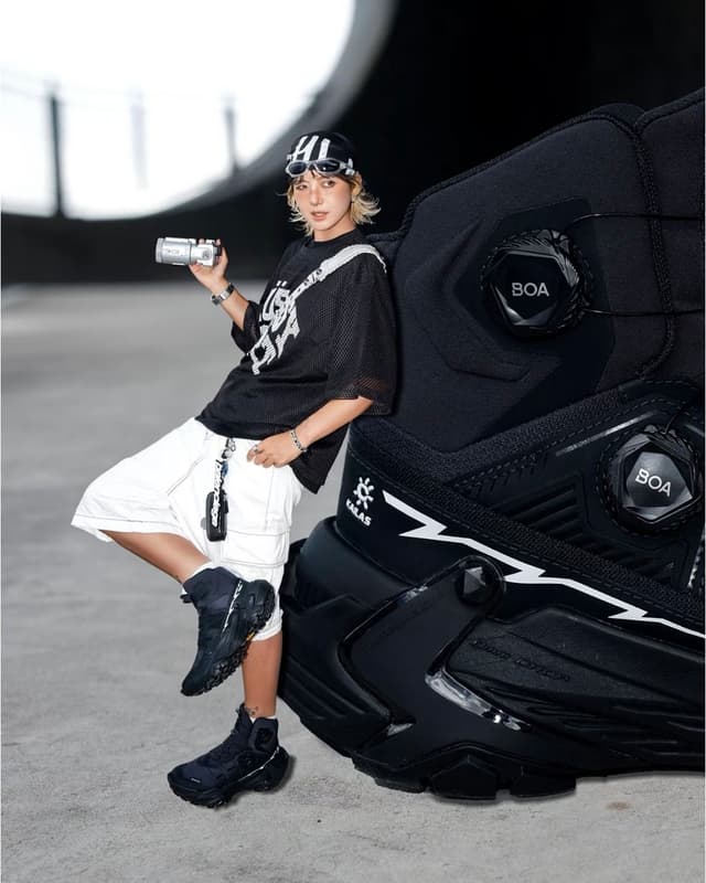 A stylish person in black and white streetwear holds a camcorder, leaning on an oversized black KALAS boot with BOA dials.