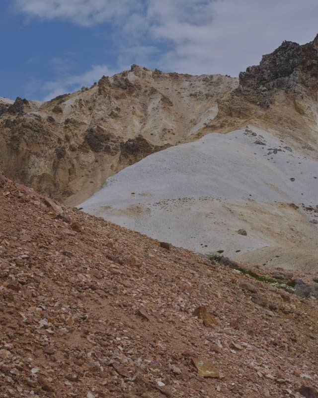 A rugged mountain scene with a reddish-brown rocky slope in the foreground leading up to mountains with tan and white rock, under a cloudy blue sky.