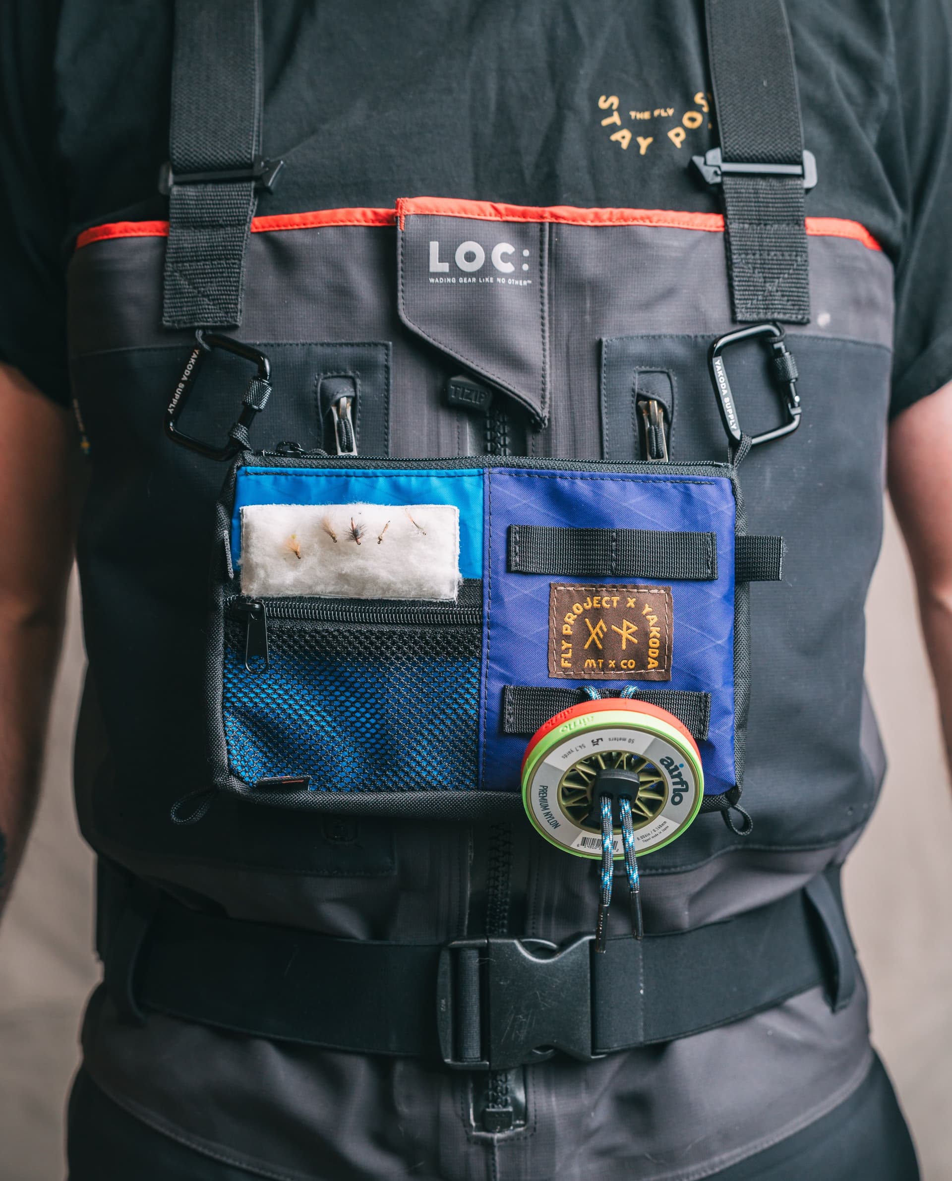 Close-up of a person wearing grey LOC fishing waders with a blue and purple chest pack holding fishing flies and an Airflo fly line spool.