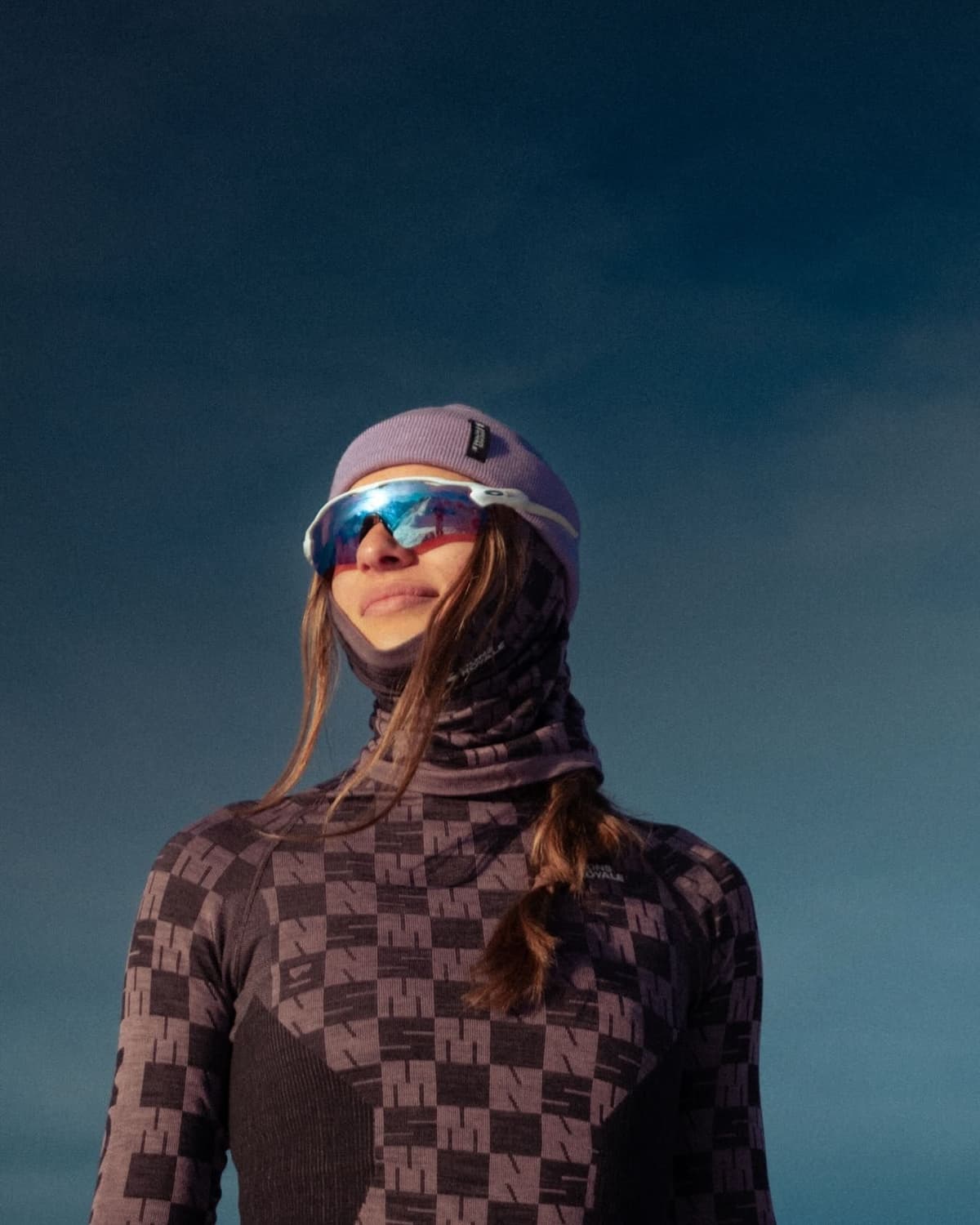 A woman in a lavender beanie, white-framed mirrored sunglasses, and a dark checkered long-sleeve shirt looks up against a blue sky.