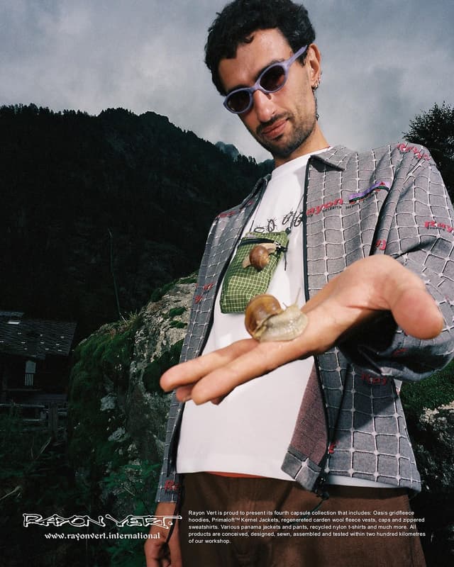 A man in purple sunglasses looks at the camera while holding a large snail on his hand, with a smaller snail on his shoulder bag, set against dark mountains.