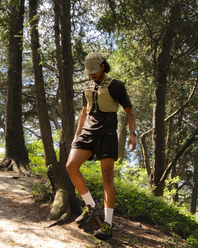 A man wearing a tan running vest, black shorts, and a camouflage cap hikes on a dirt path in a sunny forest.