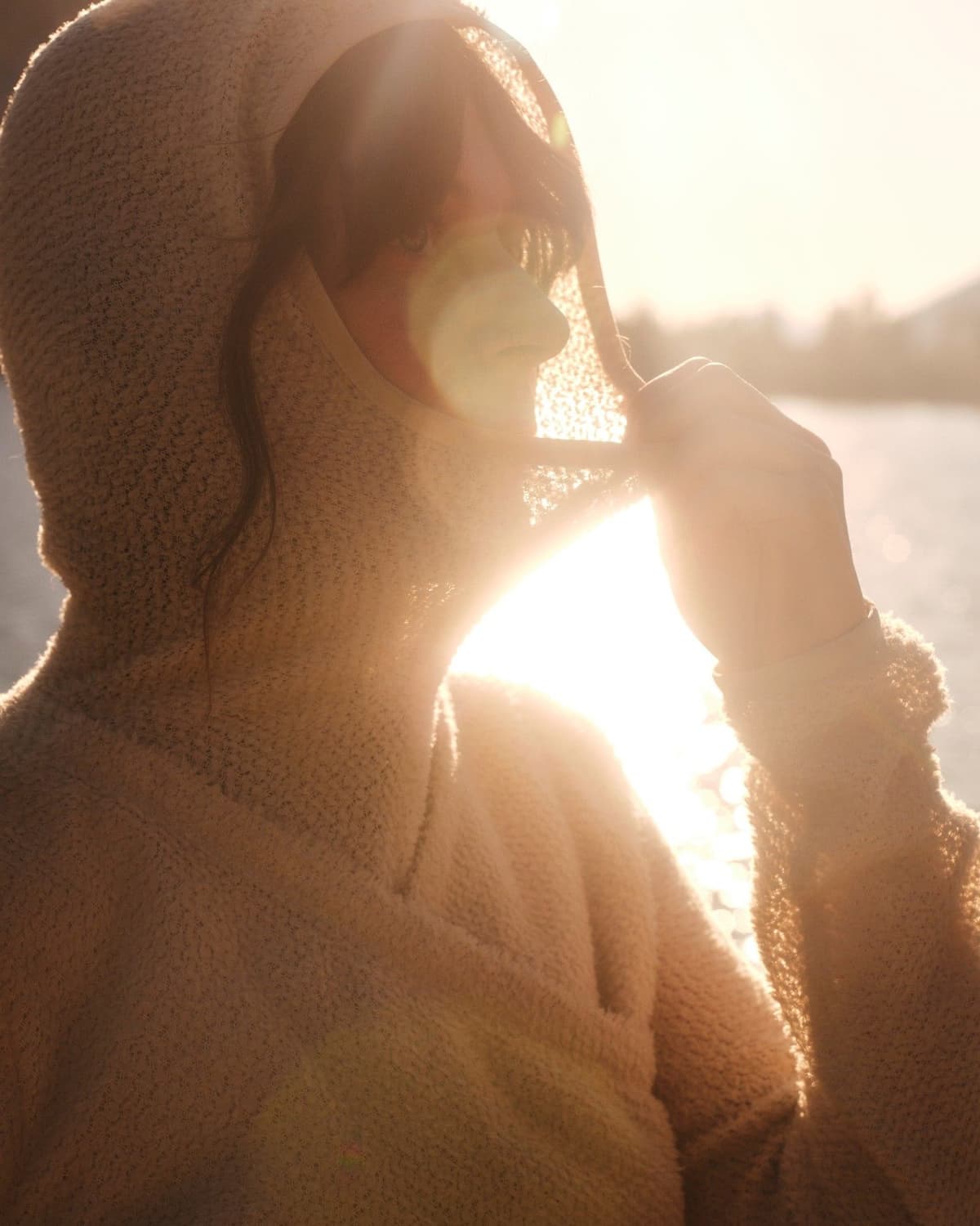 A person in a beige textured hoodie pulls the hood down, revealing part of their face, strongly backlit by golden sunlight.