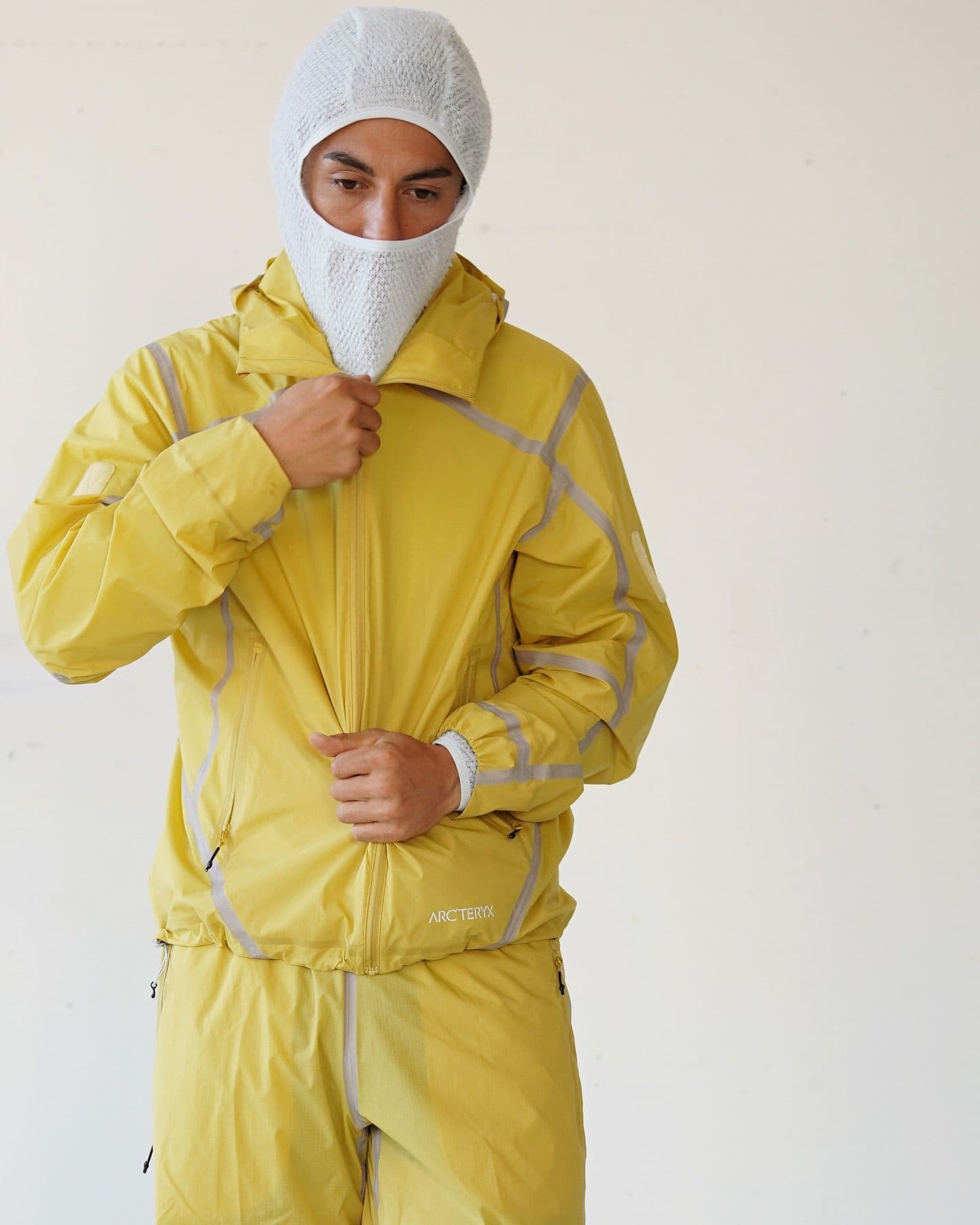 A person wearing a white mesh balaclava and a yellow Arc'teryx jacket and pants, zipping up the jacket.