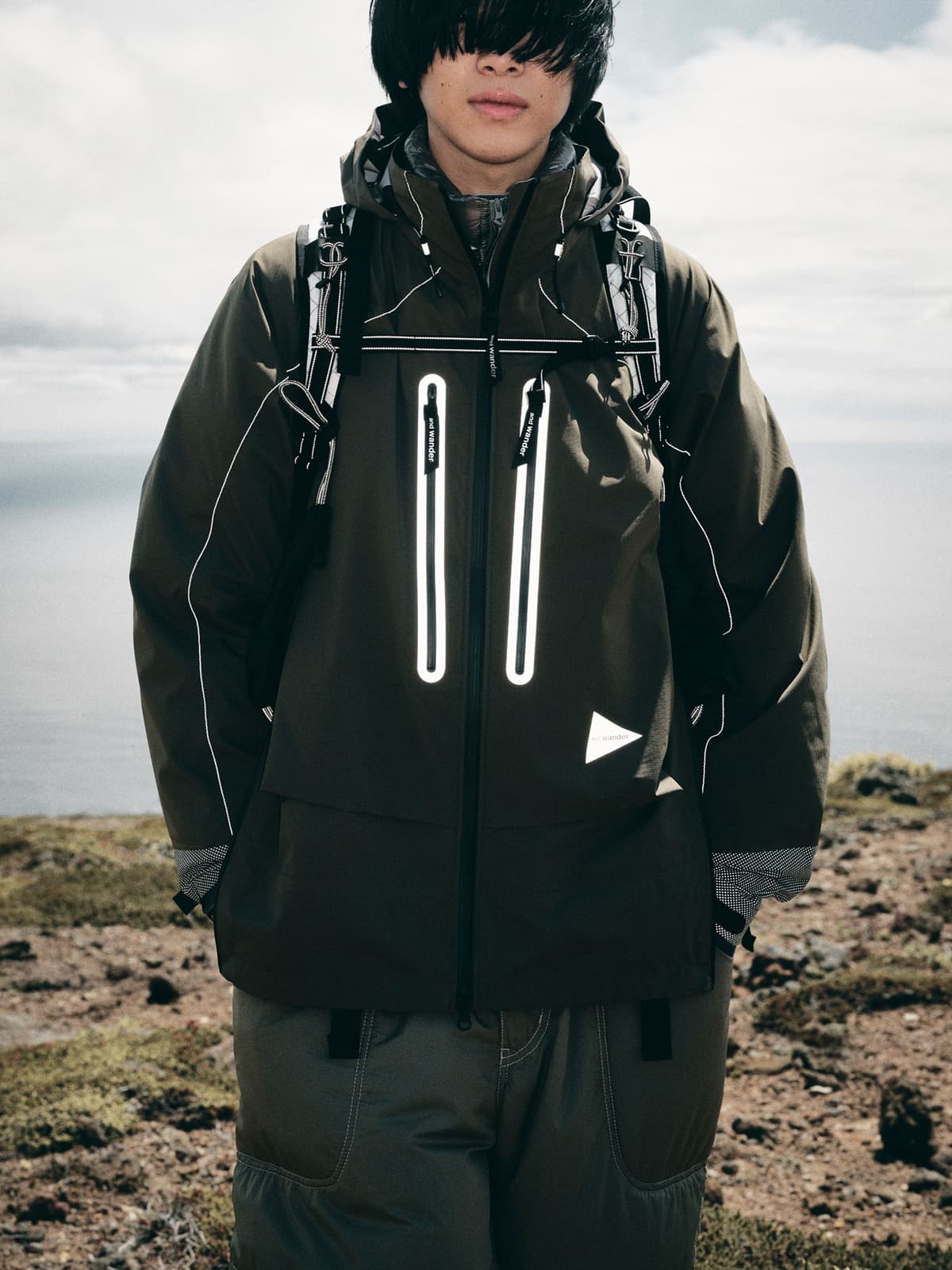 A person in a dark olive green outdoor jacket with reflective details, dark pants, and a backpack, standing on a rocky coast.