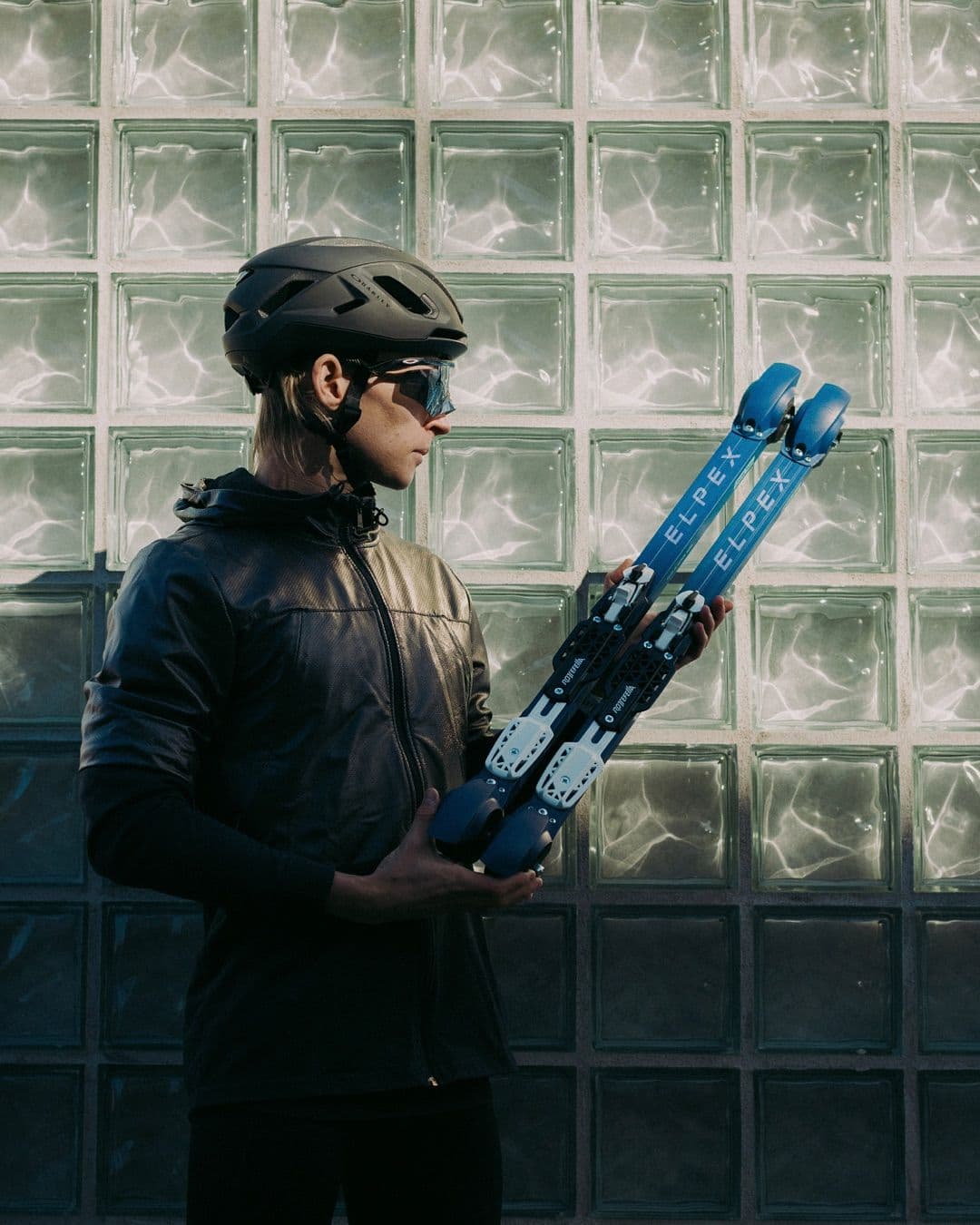 A person in a helmet and sunglasses holds blue roller skis labeled "ELPEX" against a glass block wall.
