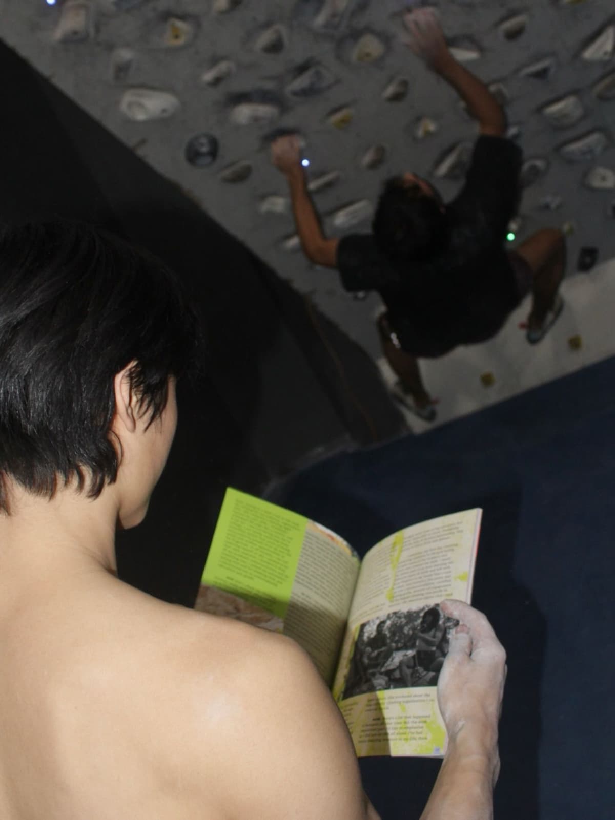 A person with chalked hands reads a book, overlooking another person climbing an indoor rock wall upside down.