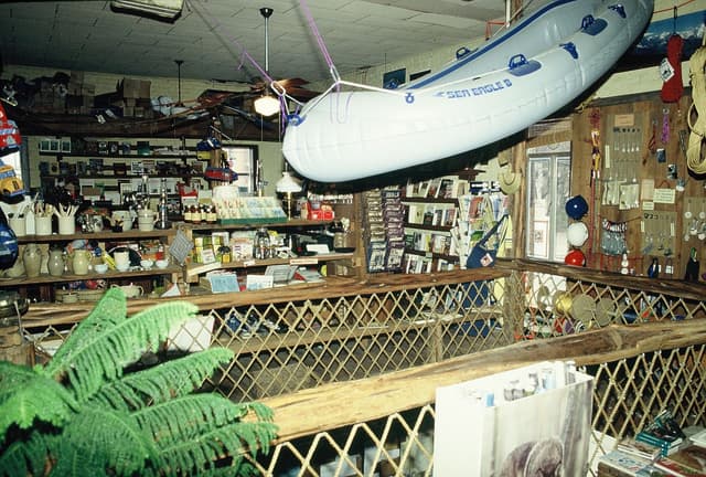 A rustic shop interior with an inflatable white raft hanging from the ceiling, packed shelves, and a wooden railing.