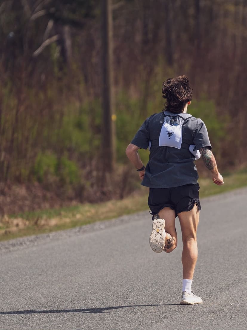 A runner seen from behind on an asphalt road, wearing a gray shirt and a light-colored hydration vest.