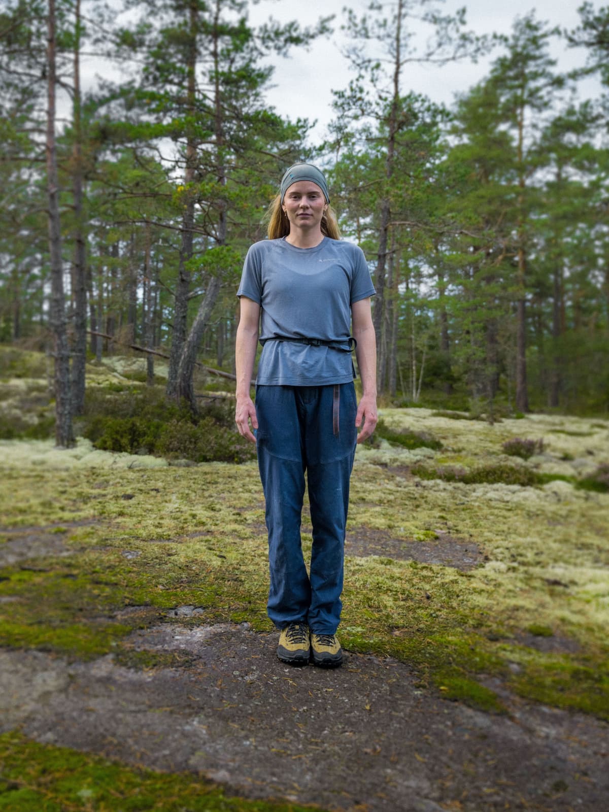 A woman in a grey shirt and blue pants stands on a mossy rock in a pine forest.