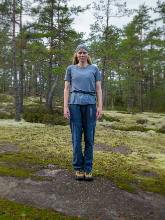 A woman wearing a gray t-shirt, blue pants, hiking shoes, and a headband stands on a mossy rock in a pine forest.