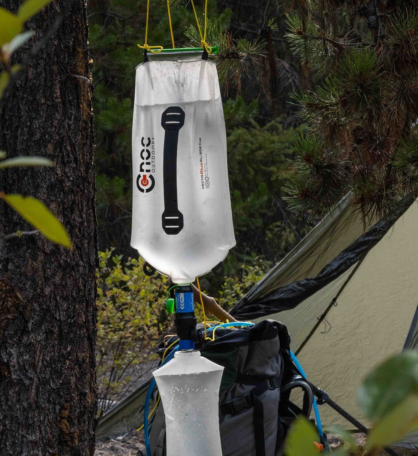 A Kroc Outdoors Vecto Dual water filter hangs from a tree, filtering water into a container at a campsite.