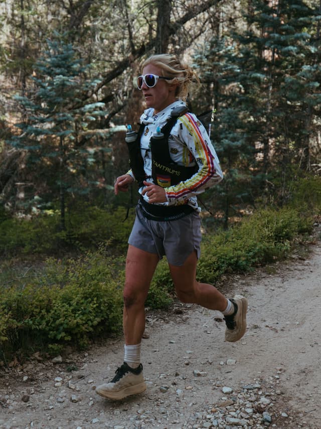 A woman in a hydration vest and sunglasses trail runs on a dirt path.