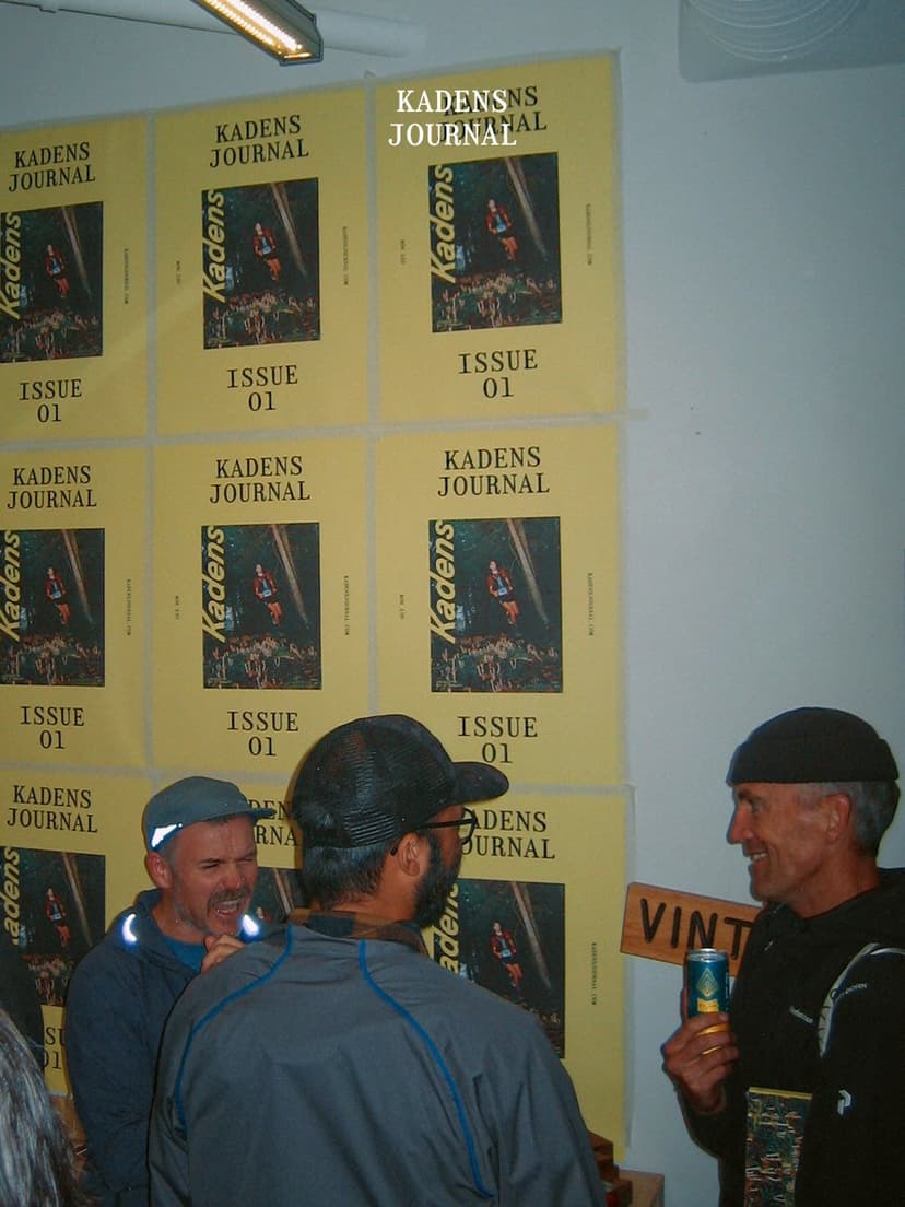 Three men talk and laugh in front of a wall covered with yellow "Kadens Journal Issue 01" posters featuring runners in a forest.